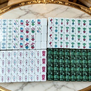 Może przedstawiać: Zestaw białych płytek do gry w Mahjong z kolorowymi ilustracjami kwiatów, ptaków, lampionów i napisem Joker, obok grupy ciemnozielonych, półprzezroczystych płytek. Płytki są ułożone w cztery prostokątne bloki na marmurowej powierzchni.