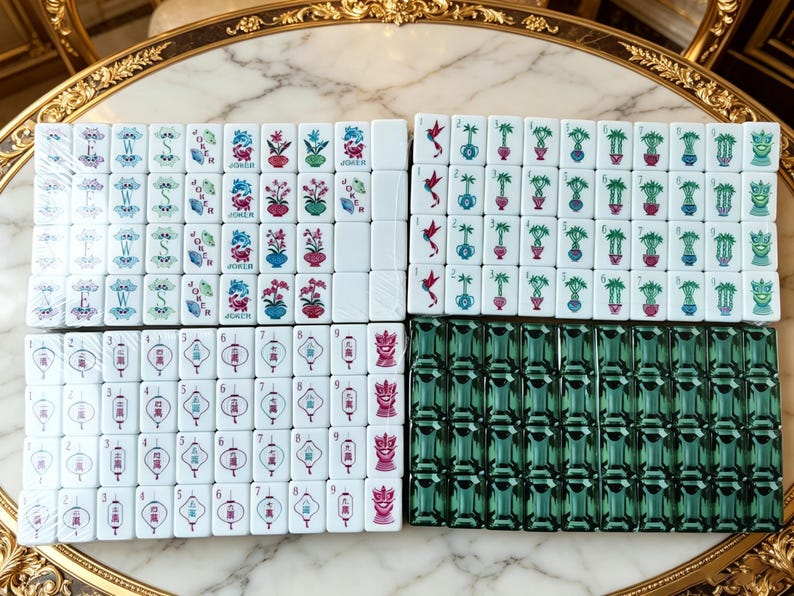 May include: A set of white Mahjong tiles featuring colorful illustrations of flowers, birds, lanterns, and the word Joker, alongside a group of solid dark green translucent tiles. The tiles are arranged in four rectangular blocks on a marble surface.