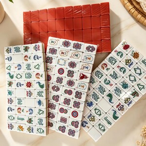 K&ouml;nnte beinhalten: Ein Mahjong-Spielset bestehend aus drei rechteckigen Anordnungen wei&szlig;er Spielsteine mit farbigen Gravuren, darunter Blumen-, Pfauen- und Joker-Motive, sowie einem Block aus einfarbig roten Spielsteinen, die in einem Raster angeordnet sind.