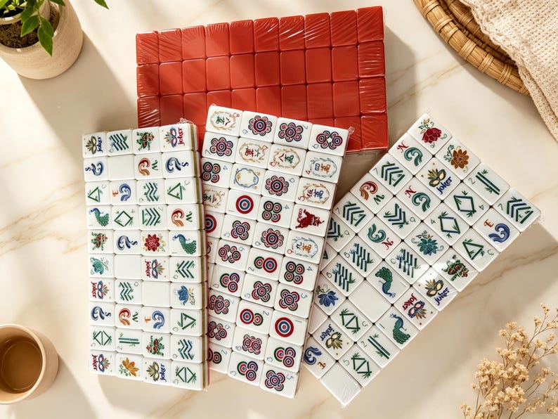 K&ouml;nnte beinhalten: Ein Mahjong-Spielset bestehend aus drei rechteckigen Anordnungen wei&szlig;er Spielsteine mit farbigen Gravuren, darunter Blumen-, Pfauen- und Joker-Motive, sowie einem Block aus einfarbig roten Spielsteinen, die in einem Raster angeordnet sind.