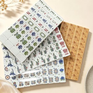 May include: A set of rectangular mahjong tiles arranged in overlapping stacks. The tiles feature white surfaces with colorful printed designs including floral patterns, bamboo stalks, directional letters, and numbers. One stack has a tan speckled finish.