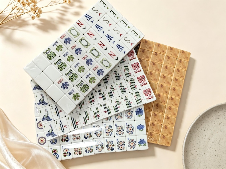 May include: A set of rectangular mahjong tiles arranged in overlapping stacks. The tiles feature white surfaces with colorful printed designs including floral patterns, bamboo stalks, directional letters, and numbers. One stack has a tan speckled finish.
