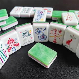 May include: A collection of rectangular mahjong tiles featuring a white base with a green marbled top layer. The tiles display various colorful illustrations including bamboo, pagodas, umbrellas, lanterns, and flowers, along with the text 'Joker'.