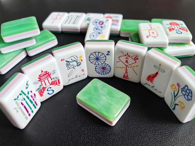 May include: A collection of rectangular mahjong tiles featuring a white base with a green marbled top layer. The tiles display various colorful illustrations including bamboo, pagodas, umbrellas, lanterns, and flowers, along with the text 'Joker'.