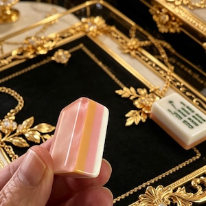 May include: A rectangular mahjong tile featuring a smooth, tri-colored surface with vertical stripes of translucent pink, solid orange, and opaque white. The tile has rounded edges and a polished finish, presented against a black velvet tray.