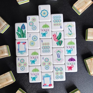Puede incluir: Una colecci&oacute;n de fichas de Mahjong blancas con ilustraciones coloridas de paraguas, monos, flores y patrones geom&eacute;tricos. Varias fichas muestran el texto Joker, mientras que otras presentan n&uacute;meros o las letras N y W con reverso de bamb&uacute;.