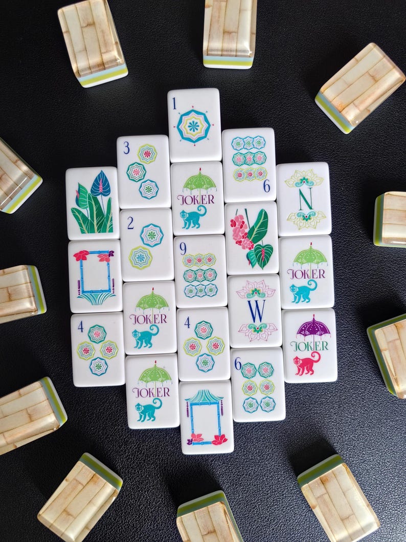Puede incluir: Una colecci&oacute;n de fichas de Mahjong blancas con ilustraciones coloridas de paraguas, monos, flores y patrones geom&eacute;tricos. Varias fichas muestran el texto Joker, mientras que otras presentan n&uacute;meros o las letras N y W con reverso de bamb&uacute;.