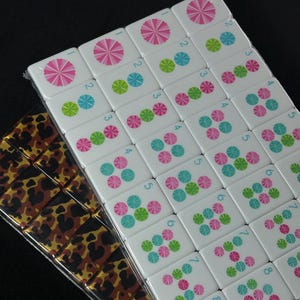 Könnte beinhalten: Ein Set rechteckiger Mahjong-Spielsteine, die in einem Raster angeordnet sind. Die Steine haben weiße Oberflächen mit rosa, blauen und grünen kreisförmigen Blumenmustern und kleinen blauen Zahlen. Ein zweites Set zeigt ein braun-schwarzes Leopardenmuster.