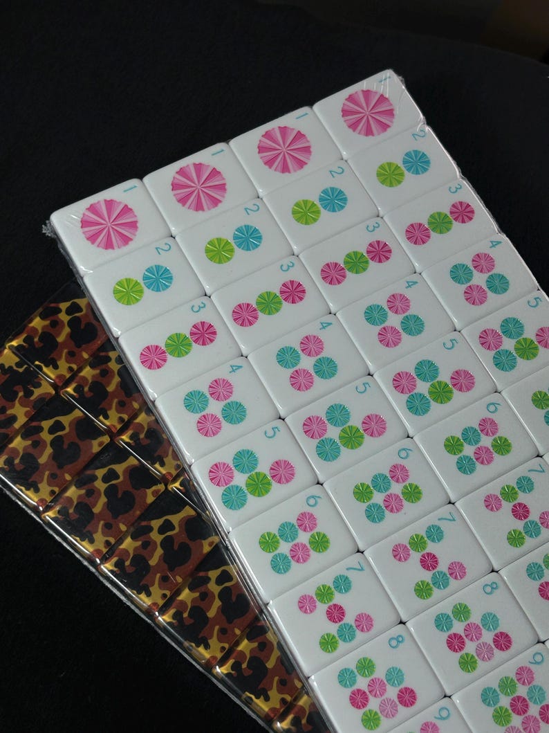 Op de afbeelding: Een set rechthoekige mahjongtegels gerangschikt in een raster. De tegels hebben witte oppervlakken met roze, blauwe en groene cirkelvormige bloemmotieven en kleine blauwe cijfers. Een tweede set tegels heeft een bruin en zwart luipaardpatroon.