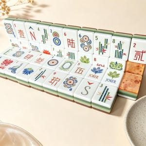 May include: A set of white Mahjong tiles arranged in two rows, featuring colorful engraved symbols, Chinese characters, numbers, and floral designs. The tiles have a green base and some display the word Joker. Two tiles on the right have a marbled texture.