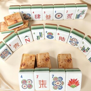 May include: A collection of rectangular mahjong tiles featuring white faces with colorful engraved symbols, numbers, and Chinese characters, paired with green and brown wood-grain patterned backs, arranged in rows and stacks on a light surface.