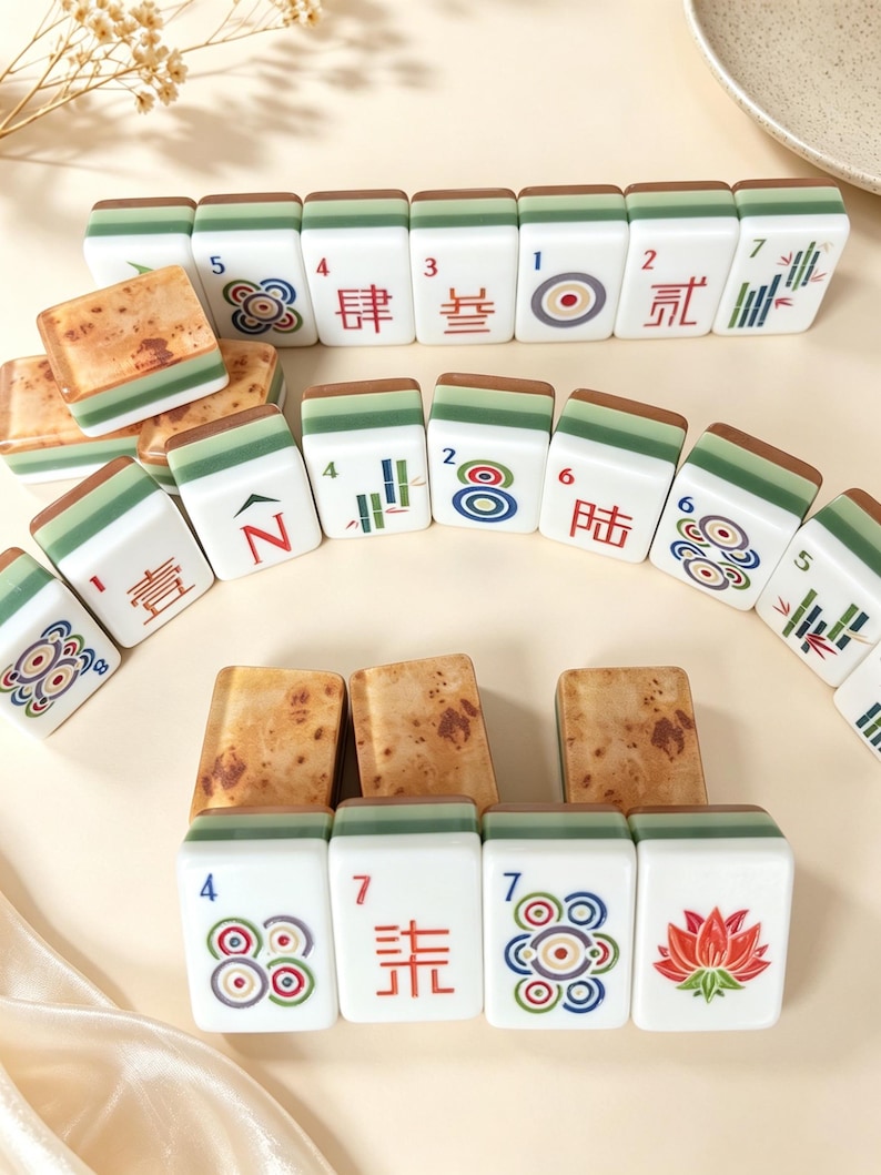 May include: A collection of rectangular mahjong tiles featuring white faces with colorful engraved symbols, numbers, and Chinese characters, paired with green and brown wood-grain patterned backs, arranged in rows and stacks on a light surface.