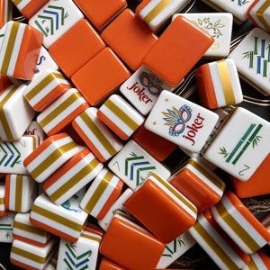 K&ouml;nnte beinhalten: Eine Sammlung rechteckiger Mahjong-Spielsteine mit einer Mischung aus einfarbigen orangefarbenen R&uuml;ckseiten und wei&szlig;en Vorderseiten. Die wei&szlig;en Fl&auml;chen zeigen verschiedene bunte Illustrationen, darunter gr&uuml;ne Bambusstangen, Blumenmuster und Joker-Steine.