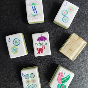 Puede incluir: Un conjunto de siete fichas de Mahjong rectangulares con caras blancas y reversos verdes. Las fichas presentan varias ilustraciones coloridas, incluyendo una letra M azul, un paraguas morado con un mono rojo y el texto JOKER, dise&ntilde;os florales y n&uacute;meros.