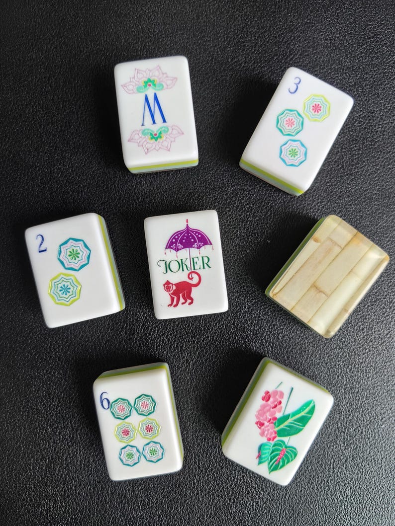Puede incluir: Un conjunto de siete fichas de Mahjong rectangulares con caras blancas y reversos verdes. Las fichas presentan varias ilustraciones coloridas, incluyendo una letra M azul, un paraguas morado con un mono rojo y el texto JOKER, dise&ntilde;os florales y n&uacute;meros.
