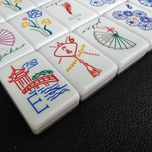 May include: A set of white rectangular mahjong tiles featuring colorful engraved illustrations including a red pagoda, a bridge, a green fan with the word joker, a red lantern, floral patterns, and blue circular designs with the number eight.