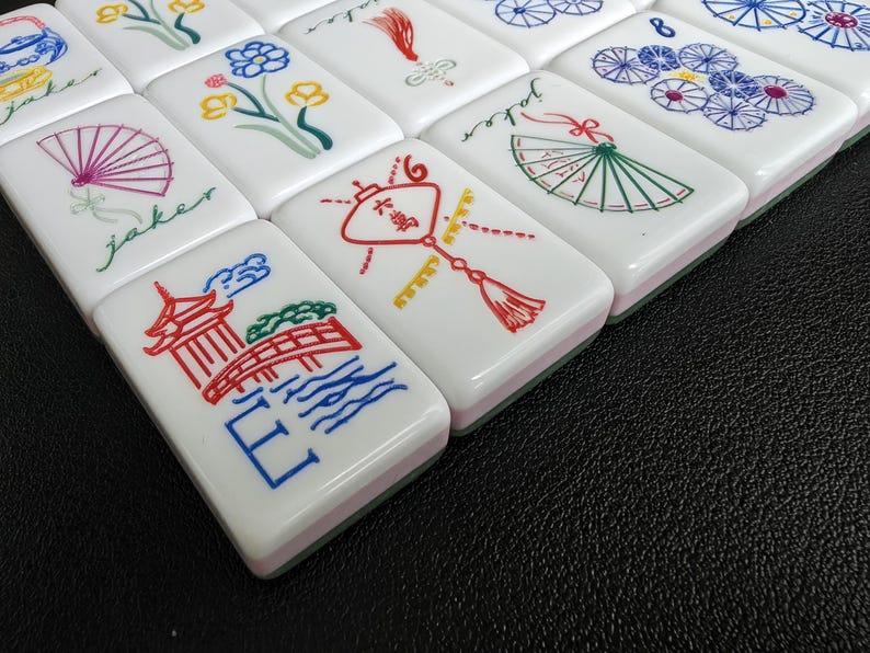 May include: A set of white rectangular mahjong tiles featuring colorful engraved illustrations including a red pagoda, a bridge, a green fan with the word joker, a red lantern, floral patterns, and blue circular designs with the number eight.