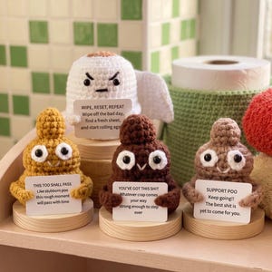 Może przedstawiać: Cztery szydełkowane figurki na drewnianych podstawkach: biała rolka papieru toaletowego z gniewną miną oraz trzy uśmiechnięte postacie w kształcie kupy w kolorze musztardowym, ciemnobrązowym i jasnobrązowym. Każda figurka trzyma białą tabliczkę.