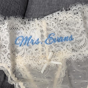 May include: Ivory lace underwear featuring a delicate scalloped edge and a small center bow. The fabric is embroidered with the text Mrs. Evans in light blue cursive script. The design includes a sheer dotted mesh panel and intricate floral patterns.