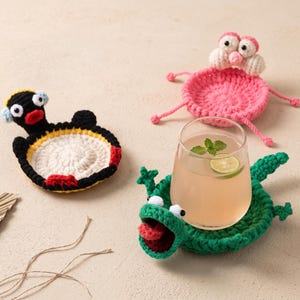 Peut inclure: Trois dessous de verre en crochet faits à la main en forme d'animaux. L'un est un pingouin noir et jaune, l'autre une créature rose aux grands yeux, et le troisième un lézard vert à la bouche ouverte contenant un verre transparent rempli.