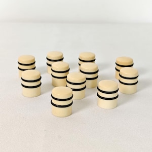 May include: A collection of ivory-colored, cylindrical objects with two black bands. The objects are arranged on a white surface. The design is simple and minimalist, with a focus on the contrast between the light and dark elements.