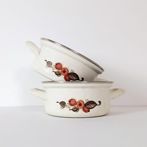 May include: Two cream-colored enamel pots with two side handles each, stacked vertically. The pots feature a painted design of red berries and brown leaves on the exterior. The rims are finished with a metallic silver-toned trim.