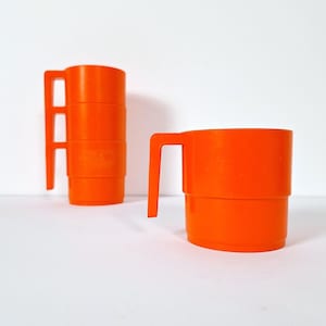 Può includere: Un set di tazze in plastica color arancione brillante dal design impilabile. Una tazza si trova da sola in primo piano, mentre una pila di tre tazze identiche è posizionata dietro sulla sinistra. Ogni tazza presenta un manico angolare piatto.