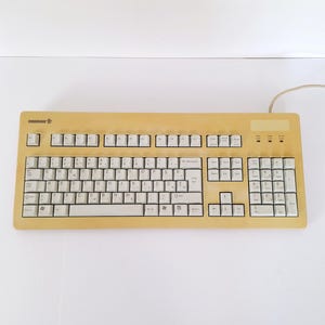 May include: A beige wired computer keyboard featuring a QWERTZ layout with white keys. The keyboard includes a numeric keypad, function keys, and a Cherry logo in the top left corner. A thin beige cable extends from the top right side of the device.