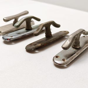 Puede incluir: Cuatro manijas de puerta de metal vintage con un diseño de palanca curvada. Las manijas tienen una apariencia envejecida y desgastada con diferentes tonos de plata y bronce. Cada manija está montada en una placa trasera rectangular con orificios para tornillos.