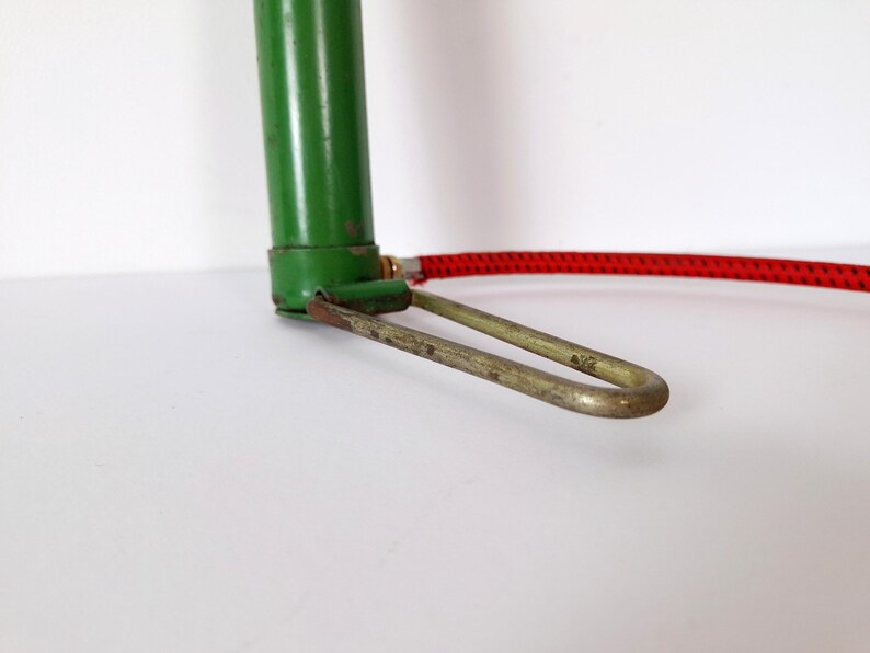 May include: Close-up of a vintage green bicycle pump with a red hose. The pump has a metal foot rest and a green cylinder. The hose is red with a black pattern.