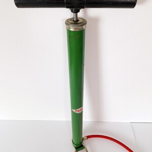 May include: A vintage bicycle pump with a black handle and a green body. The pump has a silver top and a red hose. A small label is visible on the green body. The pump is designed for inflating tires.