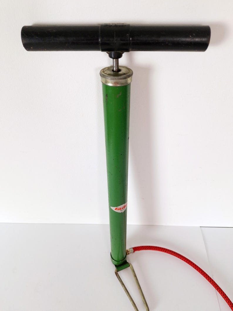 May include: A vintage bicycle pump with a black handle and a green body. The pump has a silver top and a red hose. A small label is visible on the green body. The pump is designed for inflating tires.