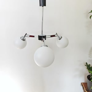 May include: A vintage ceiling light fixture with three white globe shades. The fixture has a chrome finish with black and red accents. The light is suspended from a black ceiling mount. The design is mid-century modern.