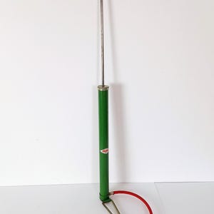 May include: A vintage bicycle pump with a black handle and a green body. The pump has a silver metal shaft and a red hose with a black nozzle. The pump is designed for inflating bicycle tires.