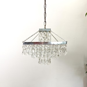 Peut inclure: Un lustre à étages doté d'un cadre carré en chrome suspendu par une chaîne en métal. Le luminaire est orné de nombreux cristaux de verre transparents et facettés, suspendus en plusieurs couches pour créer un effet de lumière en cascade.