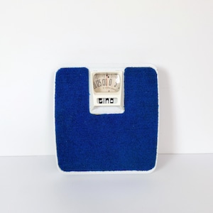 Peut inclure: Une balance de salle de bain vintage avec une surface bleue texturée et une bordure blanche. Le cadran circulaire affiche des chiffres et une aiguille. La marque "Gina" est visible.