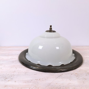 Peut inclure: Un luminaire en forme de dôme en verre blanc avec un bord marron foncé et un petit fleuron décoratif en bronze. Le luminaire repose sur une surface claire sur fond blanc.