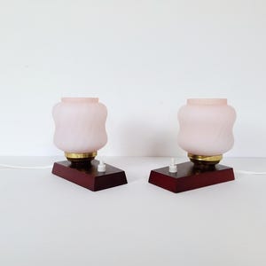 Op de afbeelding: Twee vintage tafellampen. Elke lamp heeft een roze, matglazen kap op een messing voet, gemonteerd op een donkerhouten voet. Elke lamp heeft een witte aan/uit-schakelaar en een wit snoer. De lampen staan tegen een witte achtergrond.