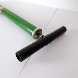 May include: A green and silver vintage bicycle pump with a black handle and a red cord. The pump is lying on a white surface. The pump is a classic design.