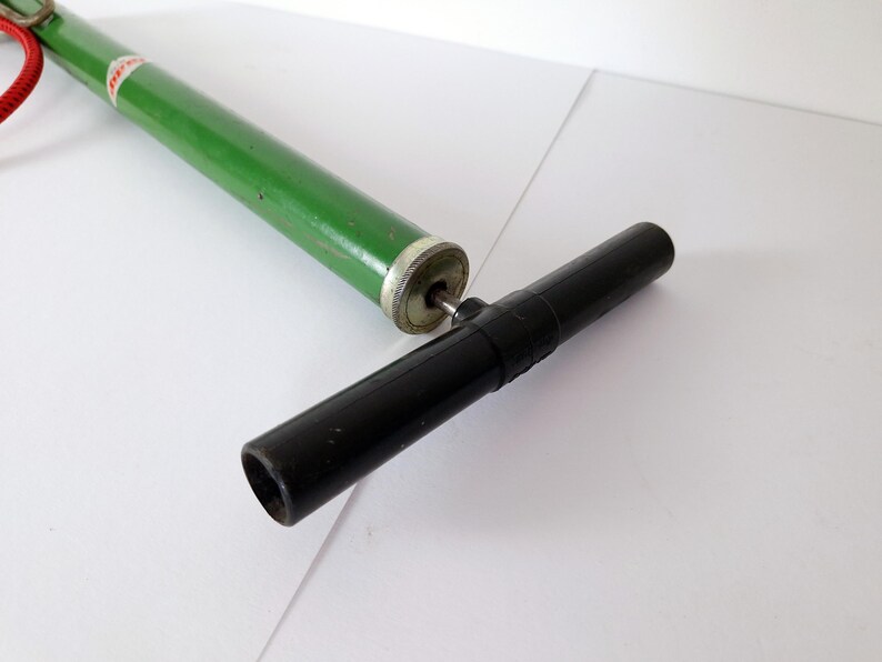May include: A green and silver vintage bicycle pump with a black handle and a red cord. The pump is lying on a white surface. The pump is a classic design.