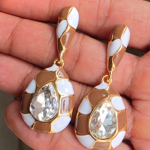 May include: A pair of drop earrings featuring a teardrop-shaped clear crystal center surrounded by a gold-tone metal frame with an enamel pattern of alternating white and tan patches. The earrings have a matching elongated top post design.