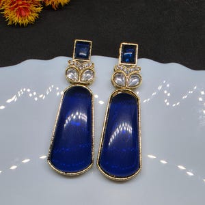 Puede incluir: Un par de pendientes colgantes dorados con un colgante rectangular grande, liso y de color azul profundo con bordes redondeados. Sobre el colgante, una pequeña piedra azul cuadrada se sitúa entre dos cristales claros en forma de gota.