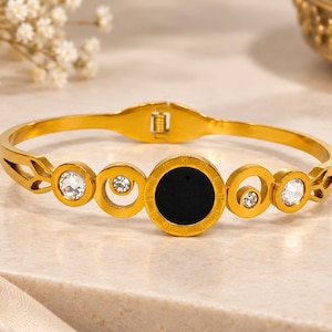 Könnte beinhalten: Ein goldfarbenes Klapparmband mit einer zentralen, kreisförmigen schwarzen Einlage, die von kleineren kreisförmigen Akzenten umgeben ist. Das Design umfasst vier klare, runde Kristalle in einem goldfarbenen Metallrahmen mit römischen Ziffern.