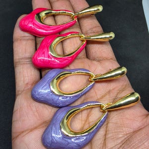 May include: Two pairs of drop earrings featuring a gold-tone metal teardrop frame attached to a smooth, irregular-shaped pendant. One pair is finished in a vibrant hot pink enamel, while the other pair is finished in a soft lavender enamel.