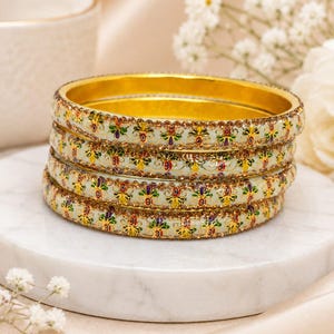 May include: A stack of four gold-toned metal bangles featuring a cream-colored enamel base. Each bangle is decorated with a repeating pattern of small, colorful floral motifs in red, blue, and green, accented by tiny, clear, sparkling rhinestones.