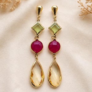 May include: A pair of gold-tone drop earrings featuring three vertically aligned stones: a light green square-cut stone at the top, a round magenta faceted stone in the centre, and a large, clear, pear-shaped faceted stone at the bottom.