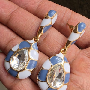 Puede incluir: Un par de pendientes colgantes con un cristal transparente en forma de lágrima en el centro, rodeado por un marco de metal dorado con segmentos de esmalte blanco y azul polvoriento. Los pendientes tienen cierre de presión y una pieza superior.