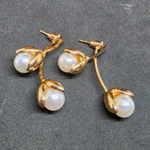 May include: A pair of gold-toned drop earrings featuring two white spherical pearls each, encased in polished gold-colored metal petals that resemble flower buds. The earrings include post-back closures and a curved stem design connecting the pearls.