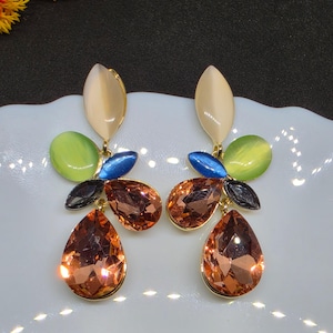 May include: A pair of dangle earrings featuring a gold-tone setting with a top cream-colored oval stone, a lime green oval stone, a small blue marquise stone, a black marquise stone, and a large faceted copper-colored teardrop crystal at the base.