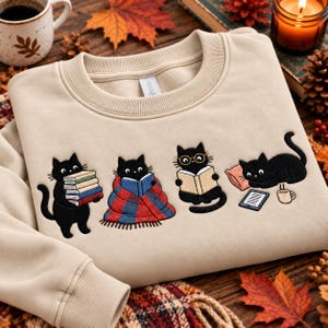 Puede incluir: Una sudadera beige de cuello redondo con un diseño bordado de cuatro gatos negros. Un gato sostiene libros, otro está envuelto en una manta de cuadros rojos y azules, uno lleva gafas mientras lee y otro está junto a una tableta y una taza.